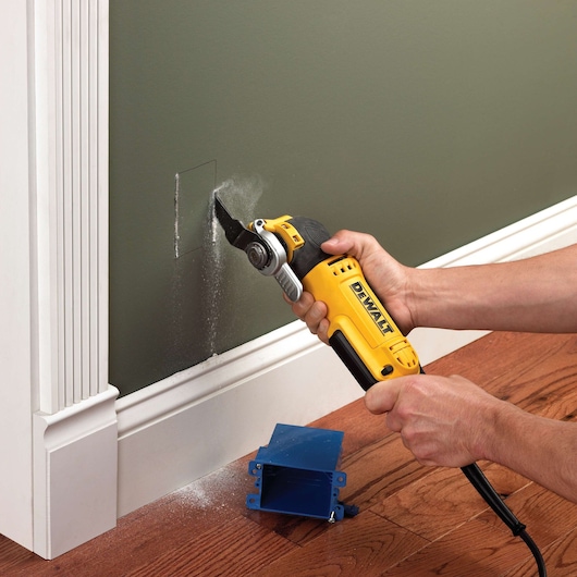 DEWALT multitool cutting a hole in a wall near a baseboard. Blue outlet box and wood floor are visible.