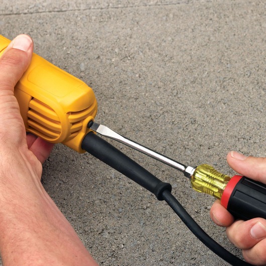 Person uses a screwdriver to adjust a cable on a yellow DEWALT power tool placed on concrete.