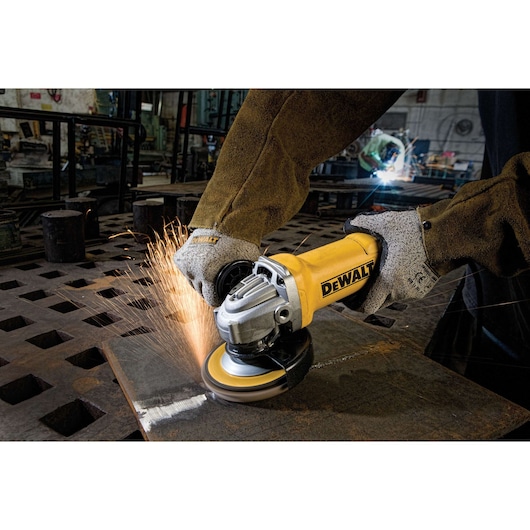 A person wearing safety gloves and a protective jacket uses a DEWALT angle grinder to cut or grind a piece of metal, producing sparks in an industrial workshop.