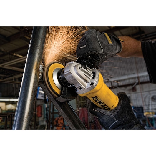 A close-up image of a DEWALT angle grinder being used on a metal pipe, with sparks flying as the tool grinds the surface. The person operating the tool is wearing protective gloves, and the scene takes place in an industrial workshop.