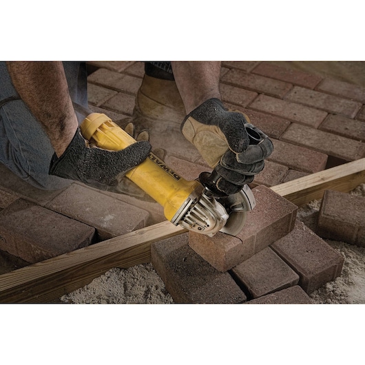A person wearing protective gloves uses a yellow DEWALT angle grinder to cut a brick paver, with dust visible, surrounded by brick pavers and a wooden beam.