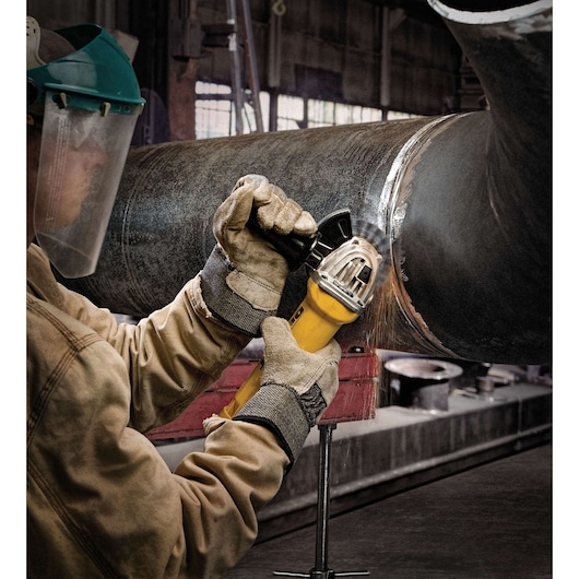 A person wearing safety gloves and a face shield is using a yellow DEWALT angle grinder to grind or cut a large metal pipe in an industrial workshop.