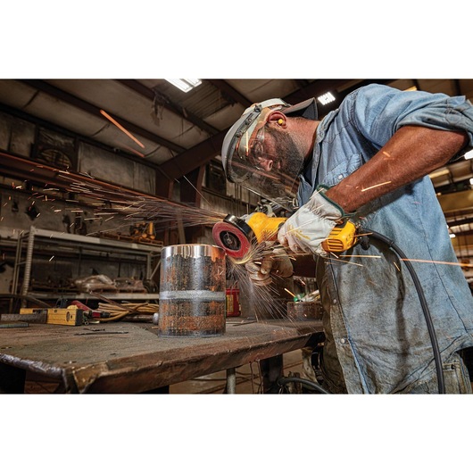 A person wearing protective gear uses a DEWALT angle grinder to grind a metal cylinder on a workbench, creating sparks in a workshop setting.