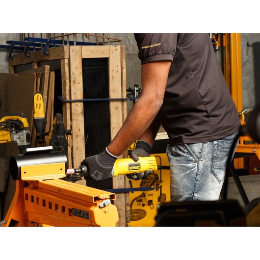 Person using a DEWALT DWE4887N die grinder on metal in a workshop with other yellow tools visible.