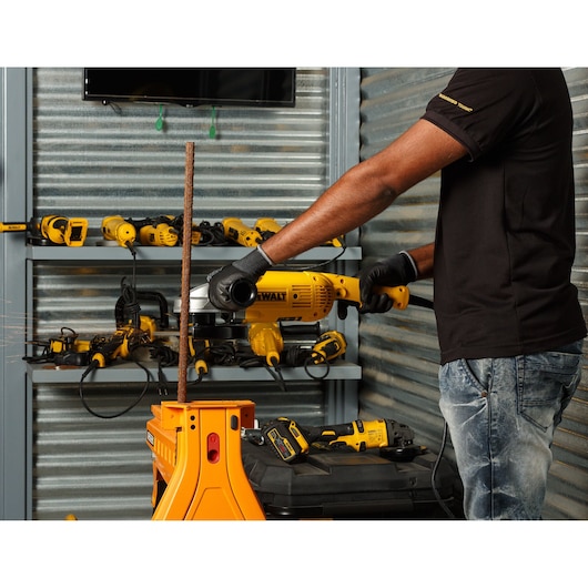 Person using DEWALT angle grinder to cut metal rod in workshop with DEWALT tools on shelves.