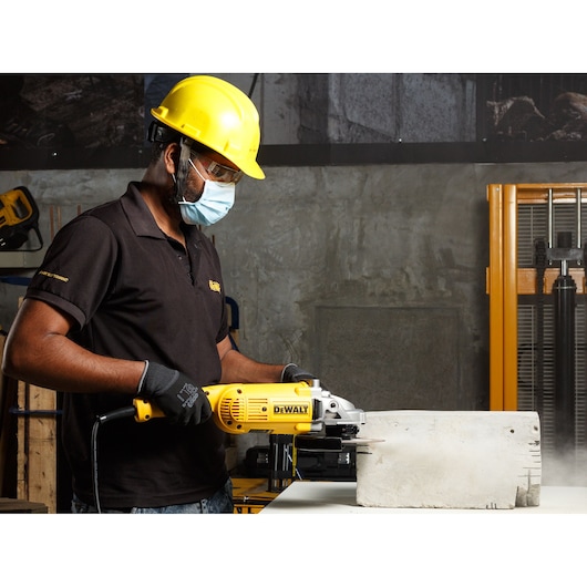 Worker using DEWALT DWE493 angle grinder to cut concrete block in industrial setting with safety gear.