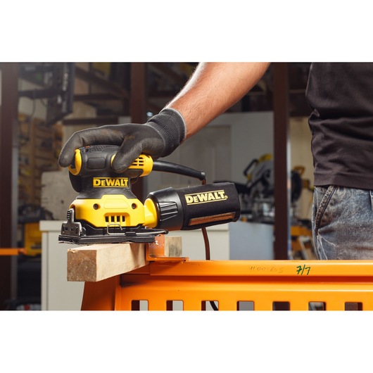 A gloved hand uses a DEWALT DWE6411 sander on a piece of wood in a workshop.