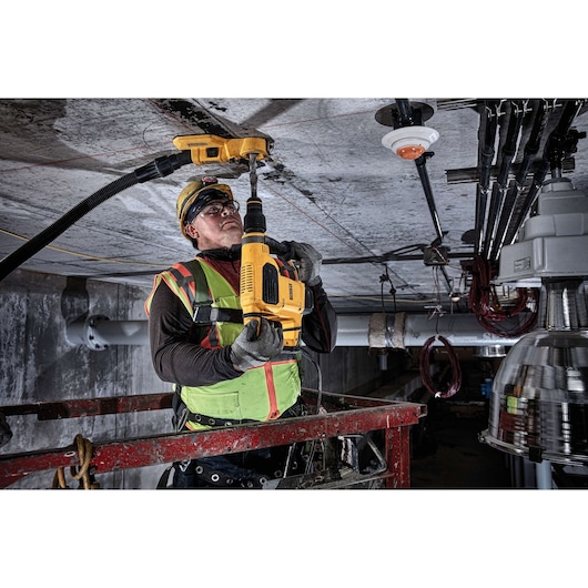 A construction worker in safety gear uses a DEWALT DWH050 tool for drilling into a ceiling in an industrial setting. The worker is standing on a lift platform, and a dust extraction attachment is connected to the tool.
