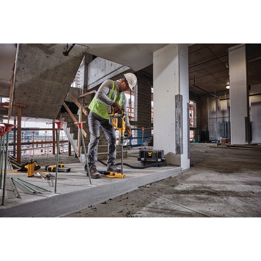 A construction worker in safety gear uses a DEWALT drilling tool with a dust extraction attachment (SKU DWH050) on a concrete floor inside an unfinished building.