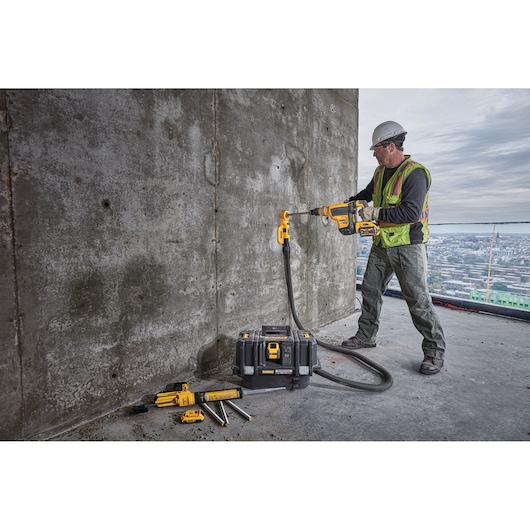 A construction worker wearing a safety vest and helmet is using a DEWALT cordless drill with dust extraction attachment to drill into a concrete wall. The worker is standing on a large building with a cityscape in the background. Several DEWALT tools and a tool case are placed on the ground nearby.