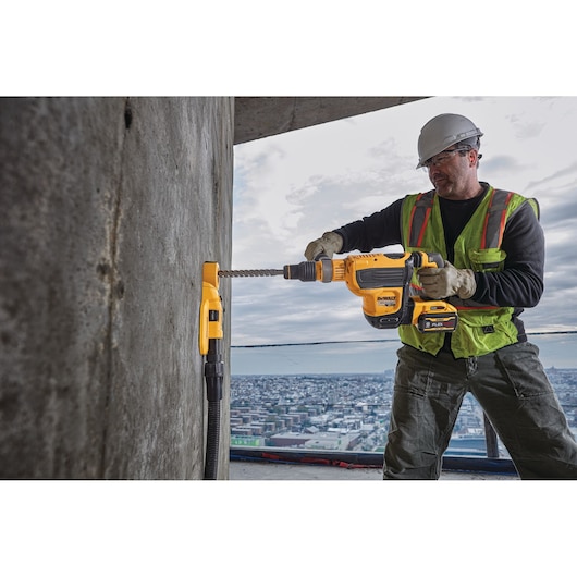 A construction worker wearing a safety helmet and vest is using a DEWALT power tool with a dust extraction attachment to drill into a concrete wall on a high-rise building site.