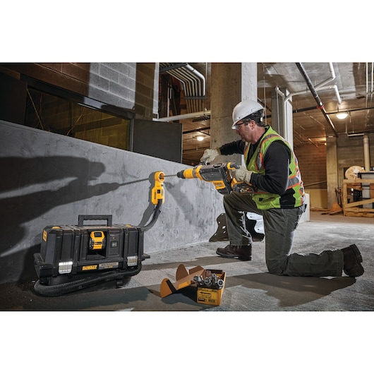 A construction worker wearing a safety vest and hard hat is using a DEWALT dust extraction system attached to a drill, working on a concrete wall in an industrial setting. The DEWALT dust extractor unit is placed on the ground nearby, with hoses connected to the drilling area. A box of drill bits is also visible beside the dust extractor.