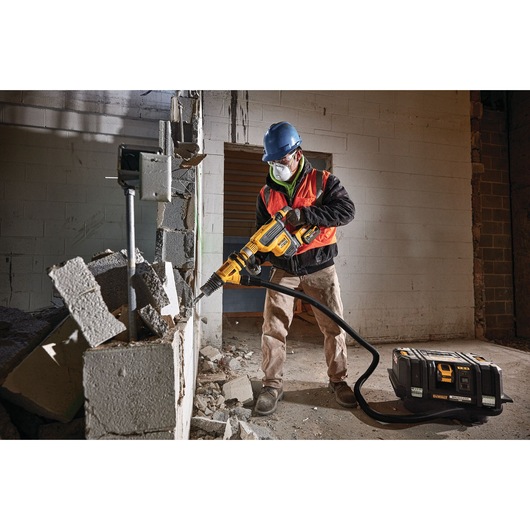 Worker using DEWALT tool to demolish a brick wall, wearing safety gear. DEWALT equipment case on floor.
