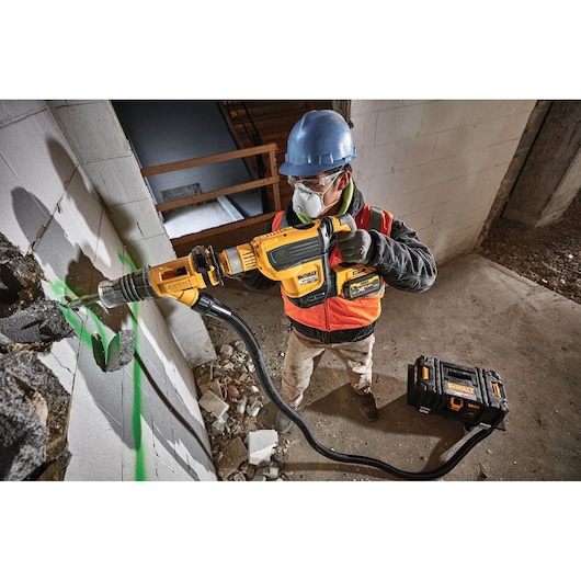 Person using a DEWALT drill with dust extractor on a concrete wall at a construction site. DEWALT toolbox nearby.