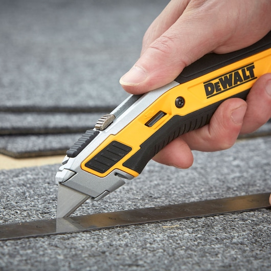 Hand uses DEWALT utility knife to cut gray carpet along a metal ruler.