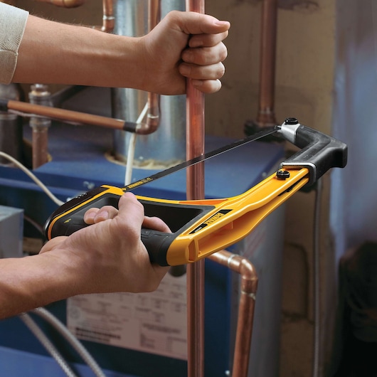 DEWALT hand saw cutting copper pipe, held by two hands in a workshop setting with visible plumbing.