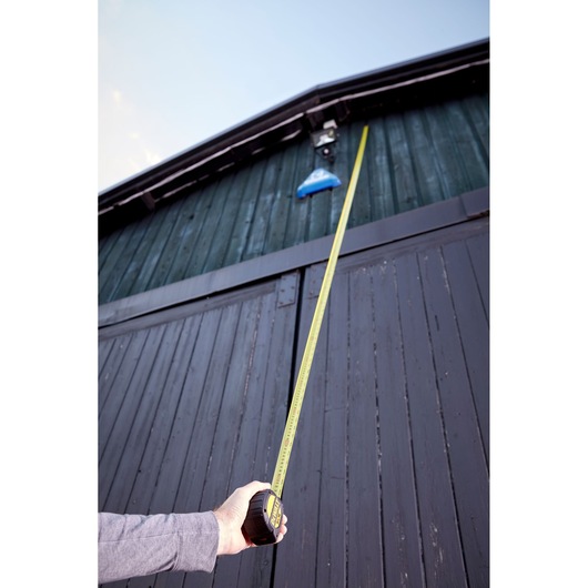 Hand holding DEWALT tape measure extended vertically against a tall wooden building exterior.