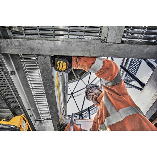 Worker in orange safety gear uses DEWALT tape measure to check distance on metal construction site.