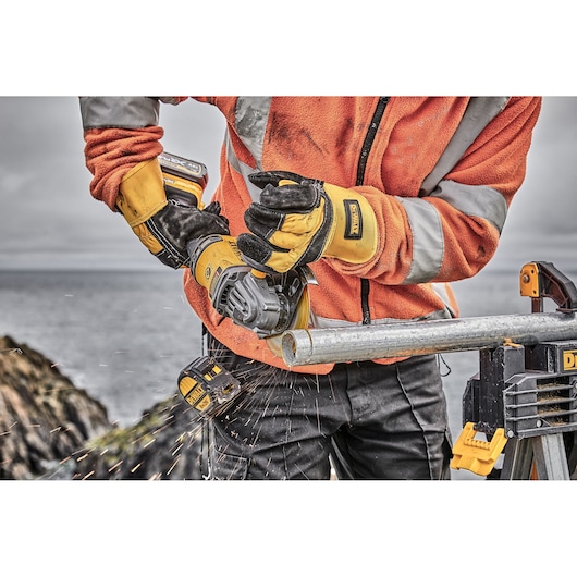Worker cuts metal pipe with DEWALT grinder, wearing safety gloves and jacket, sparks flying outdoors.