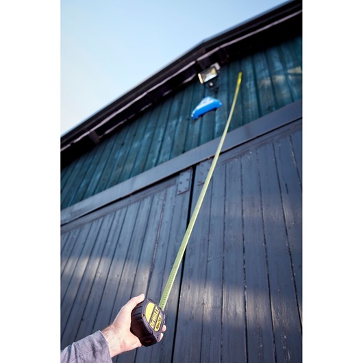 Hand using DEWALT tape measure to measure height of a tall, dark wooden building outdoors.