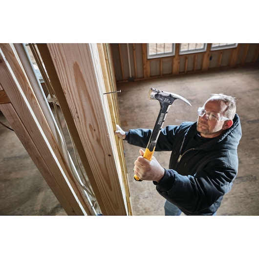 Person using a DEWALT hammer to drive a nail into wooden framing at a construction site.