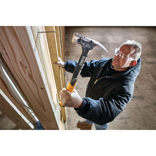 Person using a DEWALT hammer to drive a nail into wooden framing at a construction site. Face is blurred.