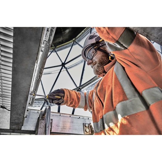 Person using a DEWALT screwdriver on metal; wearing orange safety gear and gloves at an industrial site.