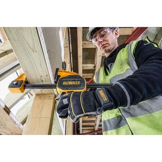 Worker uses a DEWALT clamp to secure wood during construction, wearing gloves and safety vest.