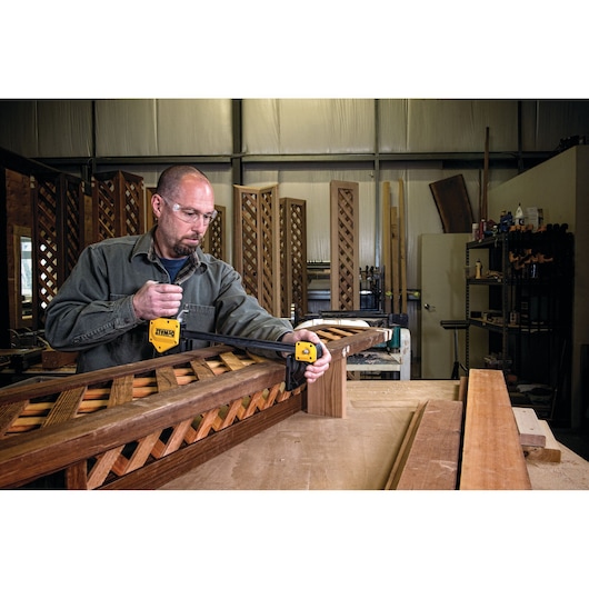 Person using a DEWALT clamp to secure wooden pieces in a workshop setting with lattice woodwork visible.