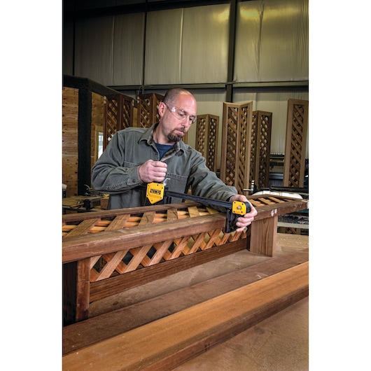 Person uses a DEWALT bar clamp on a wooden lattice in a workshop with wooden panels in the background.