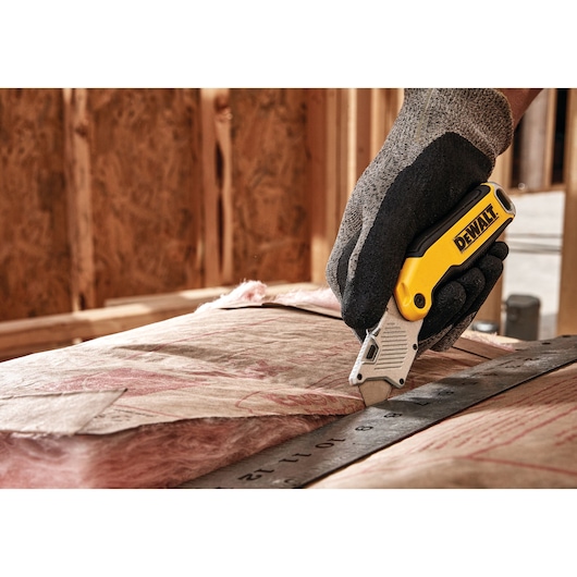 A gloved hand uses a DEWALT utility knife to cut insulation material along a metal ruler at a construction site.