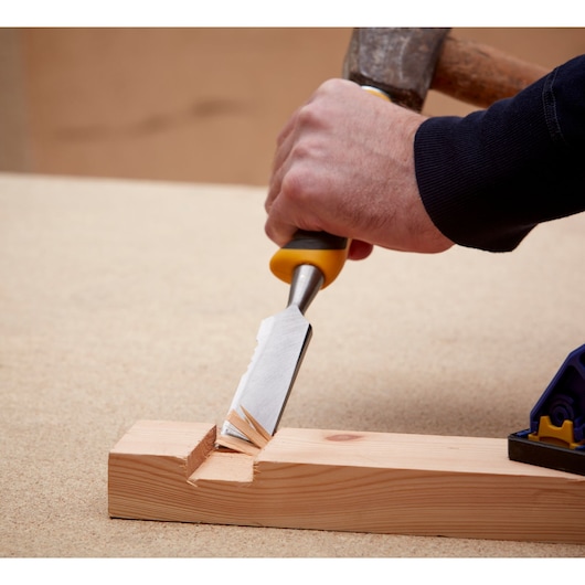 A hand uses a DEWALT chisel with yellow handle to carve wood, creating a notch in a wooden plank.