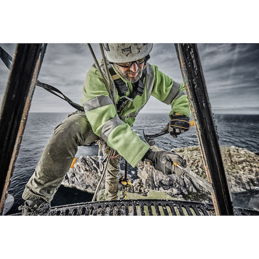 Worker uses DEWALT hammer on metal structure by the sea, wearing safety gear and green high-visibility jacket.