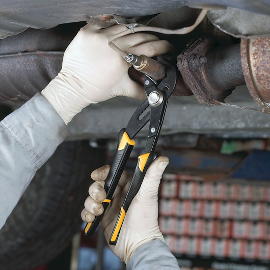 Gloved hands use DEWALT pliers to grip a metal pipe under a vehicle, performing automotive maintenance.
