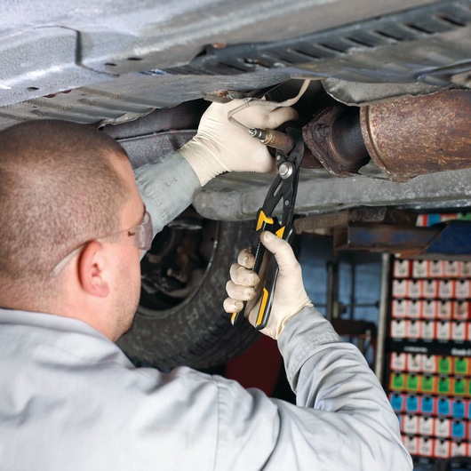 Person using DEWALT pliers to work on a car part underneath a vehicle in a garage.