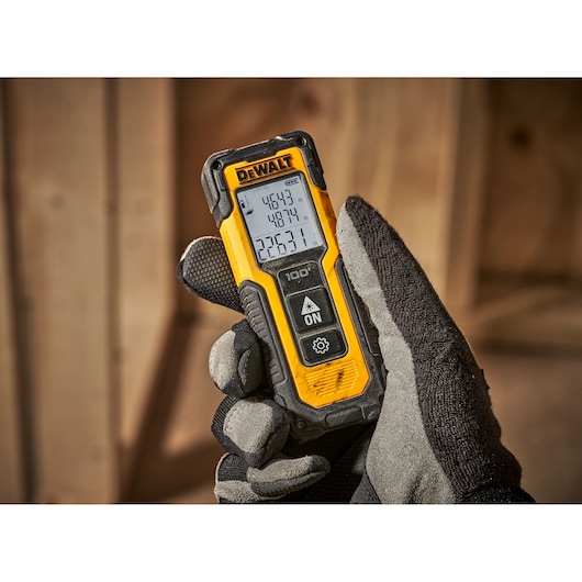 Gloved hand holding a DEWALT laser distance measuring tool with a digital display in a wooden workspace.