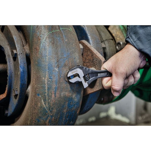 Hand using DEWALT adjustable wrench on a rusted pipe fitting in an industrial setting.