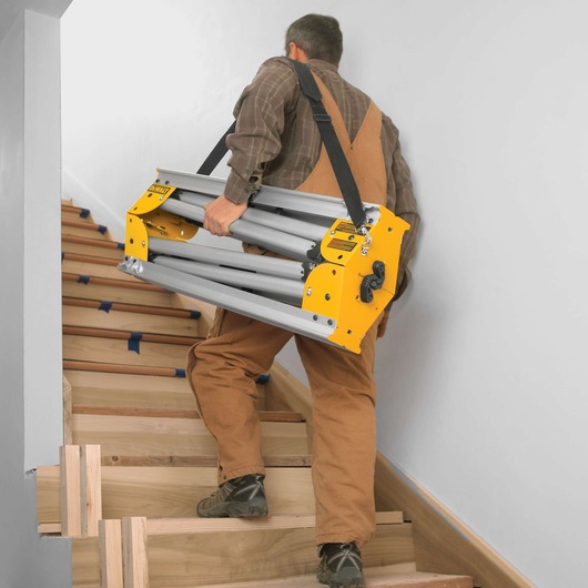 A person in work clothes carrying a folded DEWALT stand with a shoulder strap up a wooden staircase.