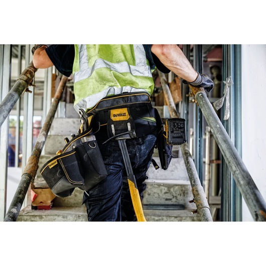 Person with DEWALT tool belt and tools, ascending a construction site staircase in workwear and safety vest.