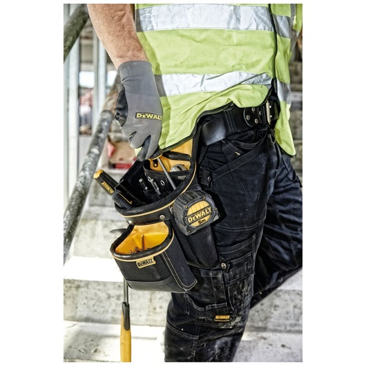 Person with DEWALT tool belt and gloves, holding tools and tape measure at a construction site.