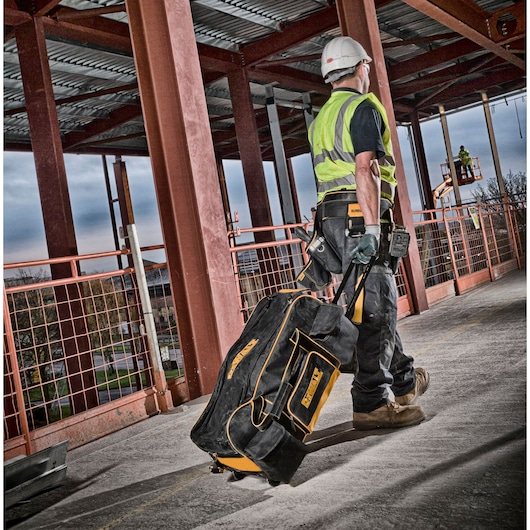 Worker pulls DEWALT rolling tool bag on construction site with steel beams and safety gear visible.