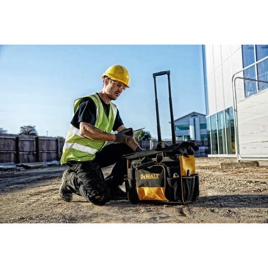 Worker with DEWALT rolling tool bag DWST1-81060 at construction site, wearing safety gear, kneeling on ground.