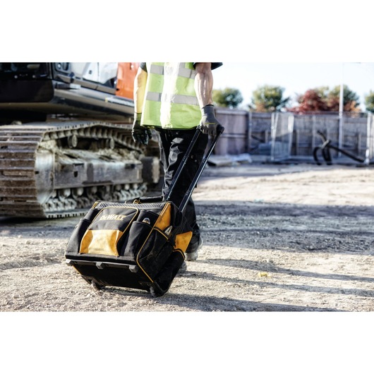 Person pulls a DEWALT rolling tool bag on a construction site, with machinery and dirt visible around.