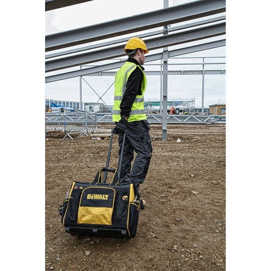 Worker pulls a DEWALT rolling tool bag at a construction site, wearing a yellow vest and helmet.
