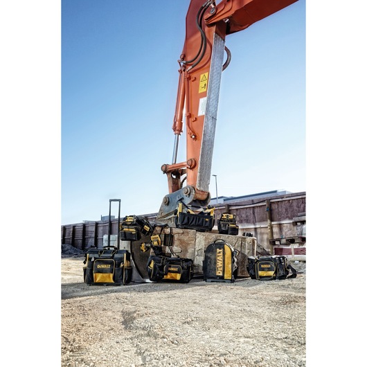 DEWALT tool bags are displayed outdoors, arranged around an excavator arm at a construction site.
