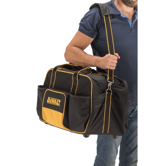 Person carrying a DEWALT black and yellow tool bag with shoulder strap, featuring pockets and wheels.