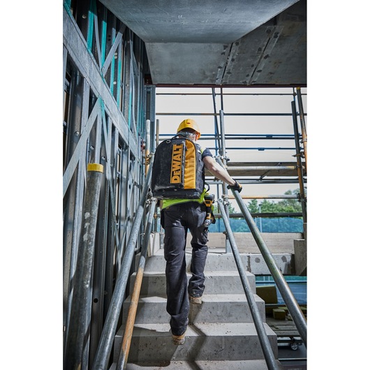 Worker with DEWALT backpack climbing stairs at a construction site, wearing a yellow helmet and safety gear.