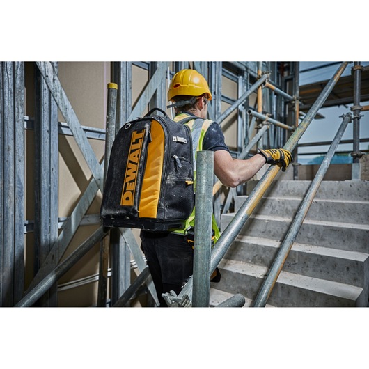 Worker with DEWALT backpack ascending stairs on a construction site, wearing safety gear and helmet.